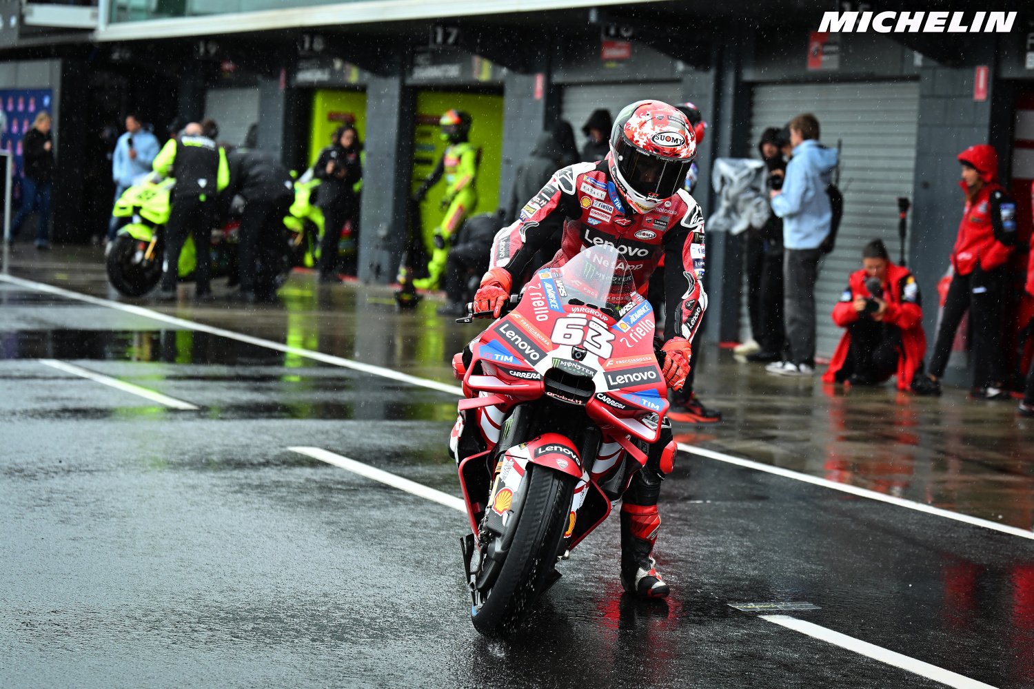 A rider in red and black leathers pushes a red Ducati MotoGP bike through a wet pit lane, race number 63 visible, Lenovo livery.