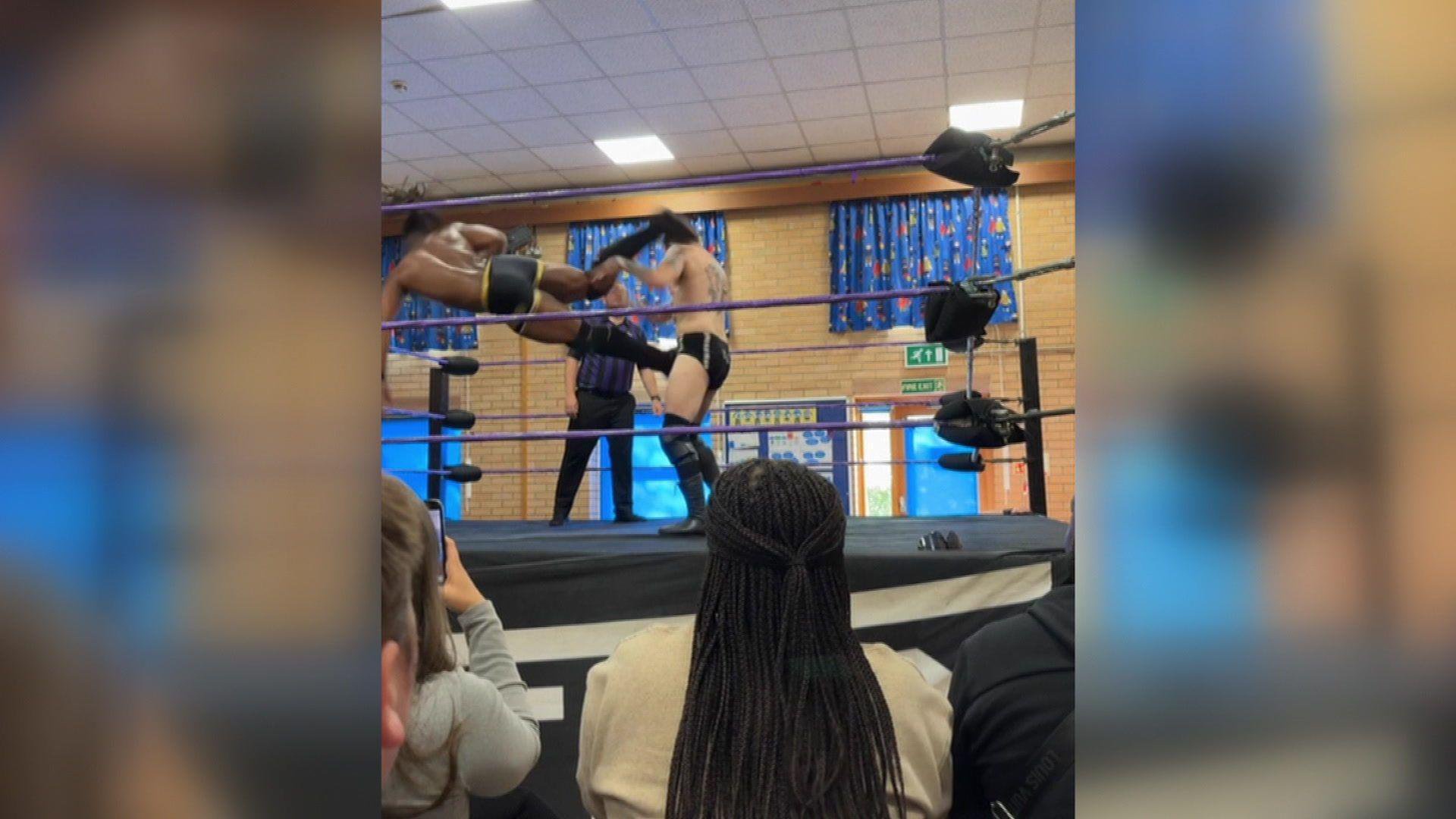 This is an action shot from a wrestling match. A man is flying through the air as he kicks his opponent. A small crowd watches on.