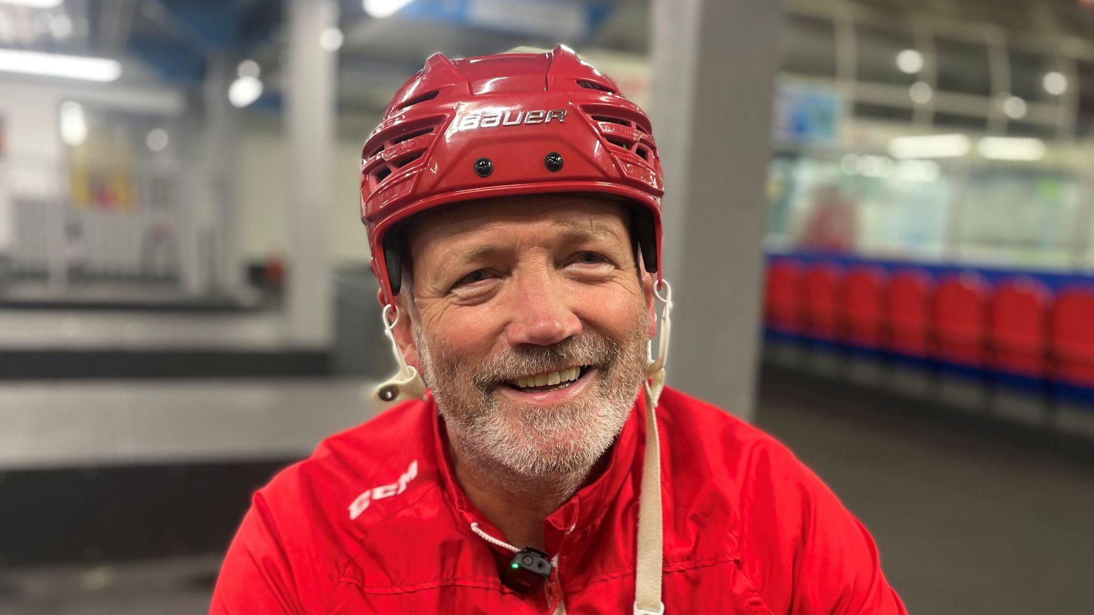 Image of a man wearing a red top, sitting on the edge of an ice rink and smiling at the camera