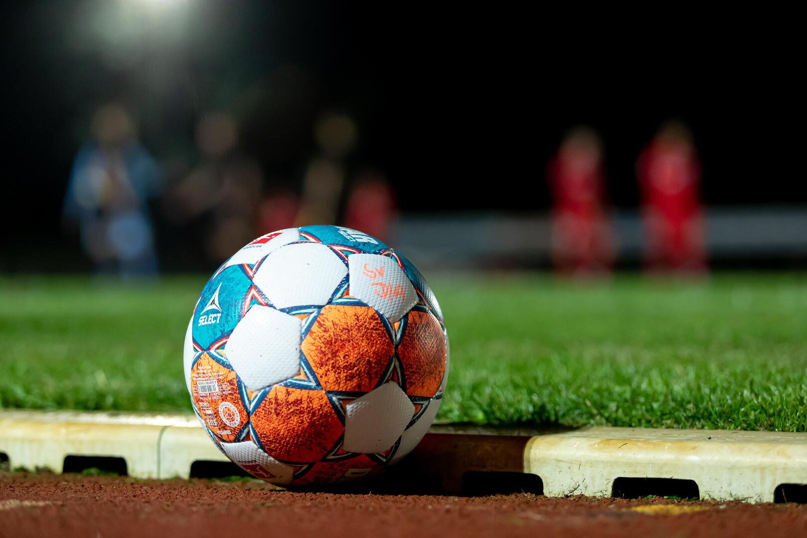 Trainingsball in Weiß, Blau und Orange. Im Hintergrund trainieren Spieler bei Flutlicht