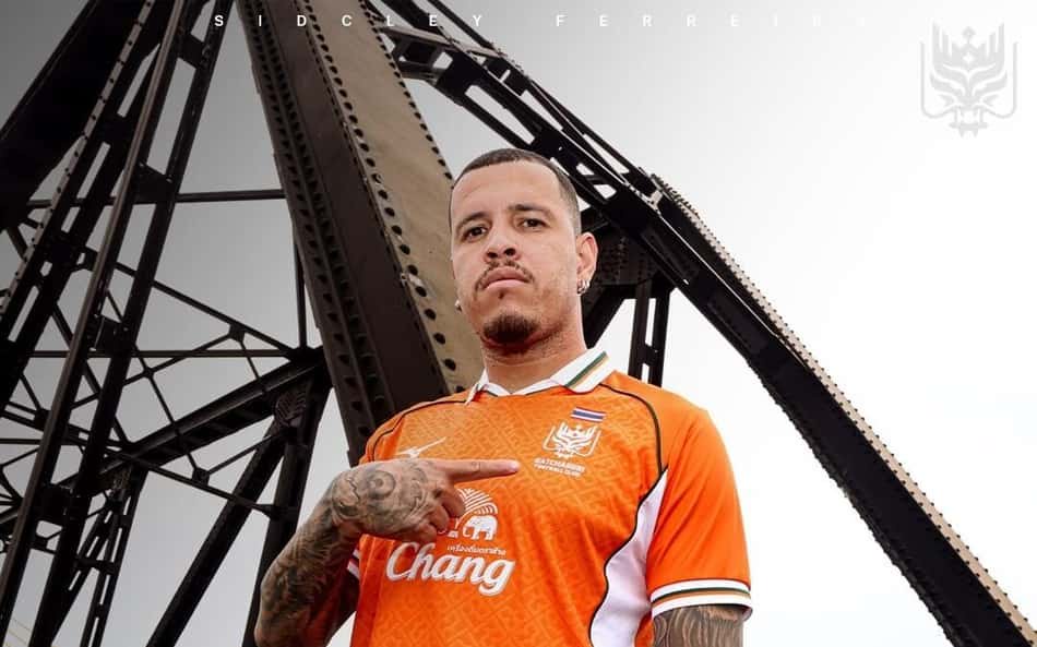 Jogador de futebol com camisa laranja do Chiangrai United, patrocinada pela Chang, apontando para o peito.
