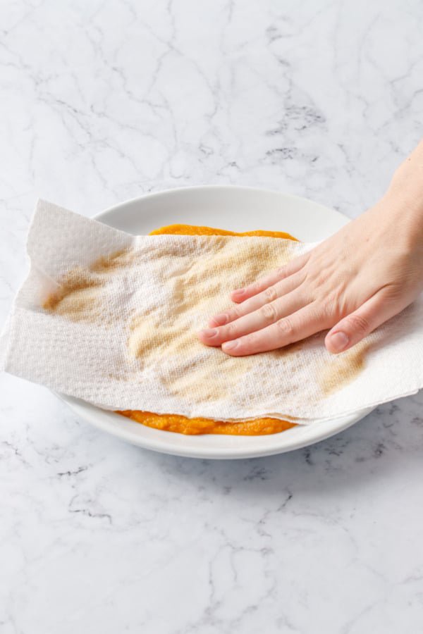 Pressing paper towels onto pumpkin puree spread on a plate to remove moisture.