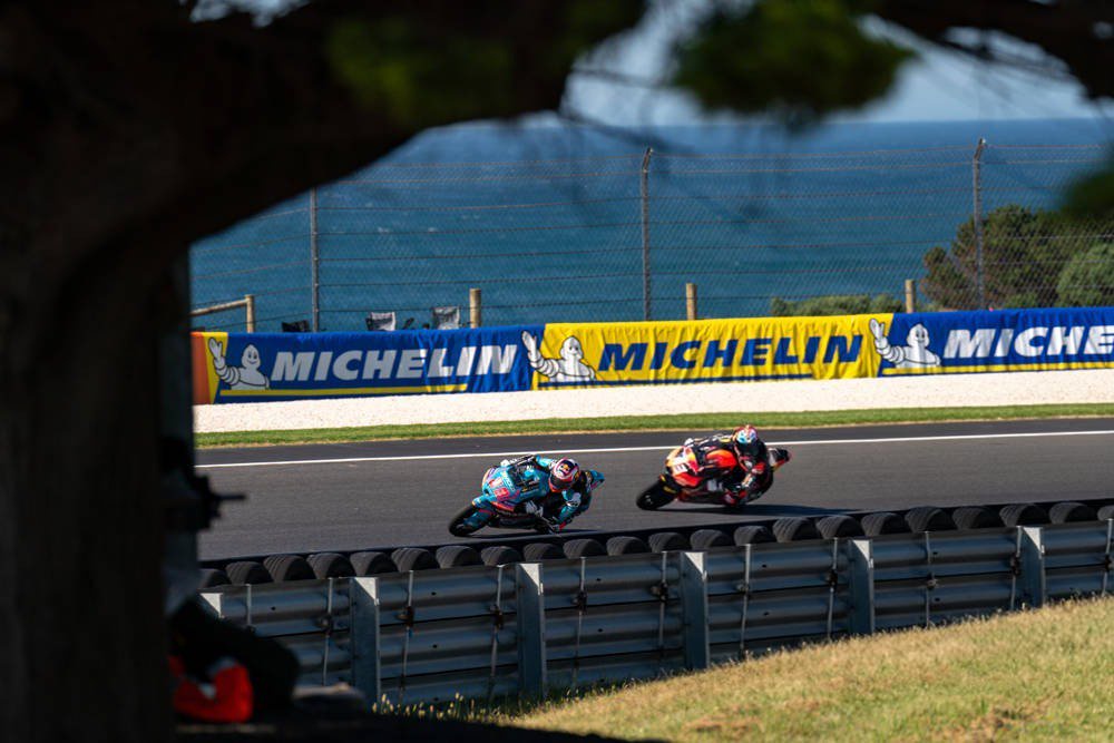 Two Moto3 riders lean into a corner at Phillip Island, one in teal and blue, one in orange and red, with Michelin barriers behind them.