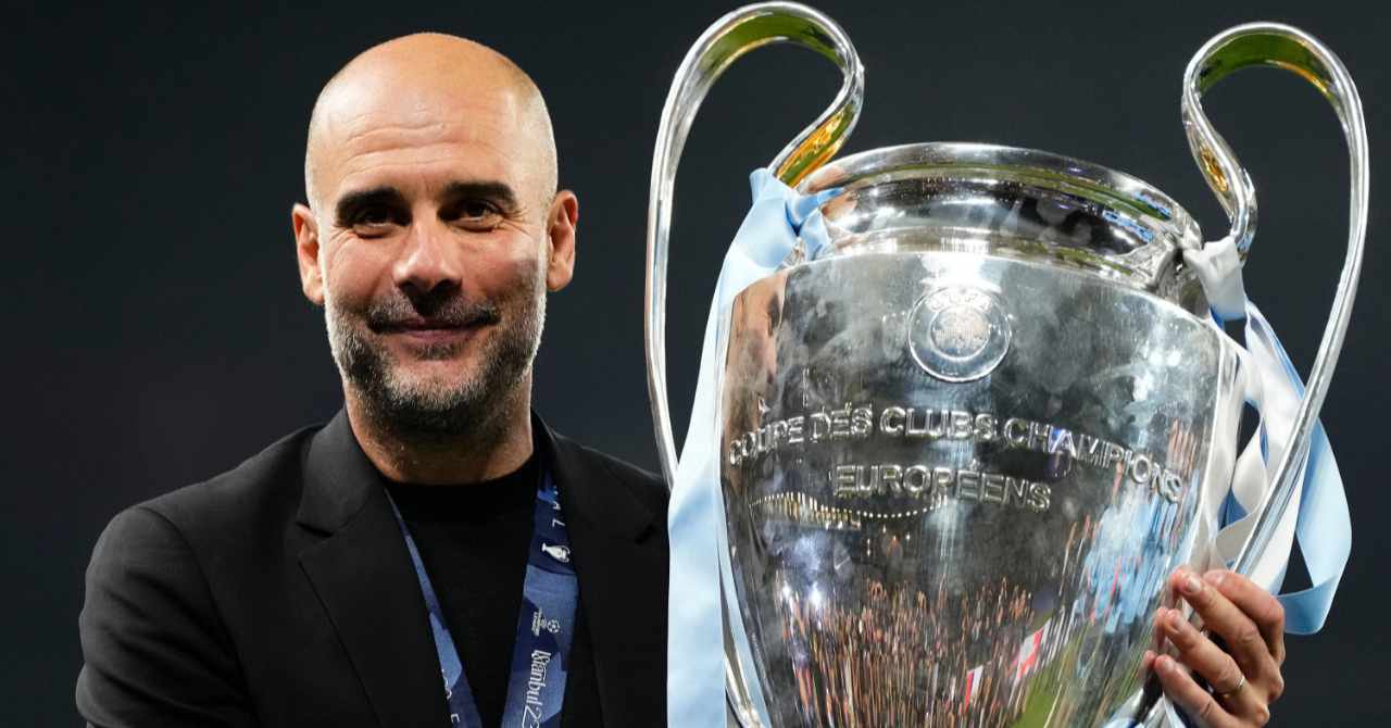 Football manager Pep Guardiola wearing a black suit while holding the silver UEFA Champions League trophy won by Man City in a stadium and smiling in his role as head coach of Premier League team Manchester City Football Club.