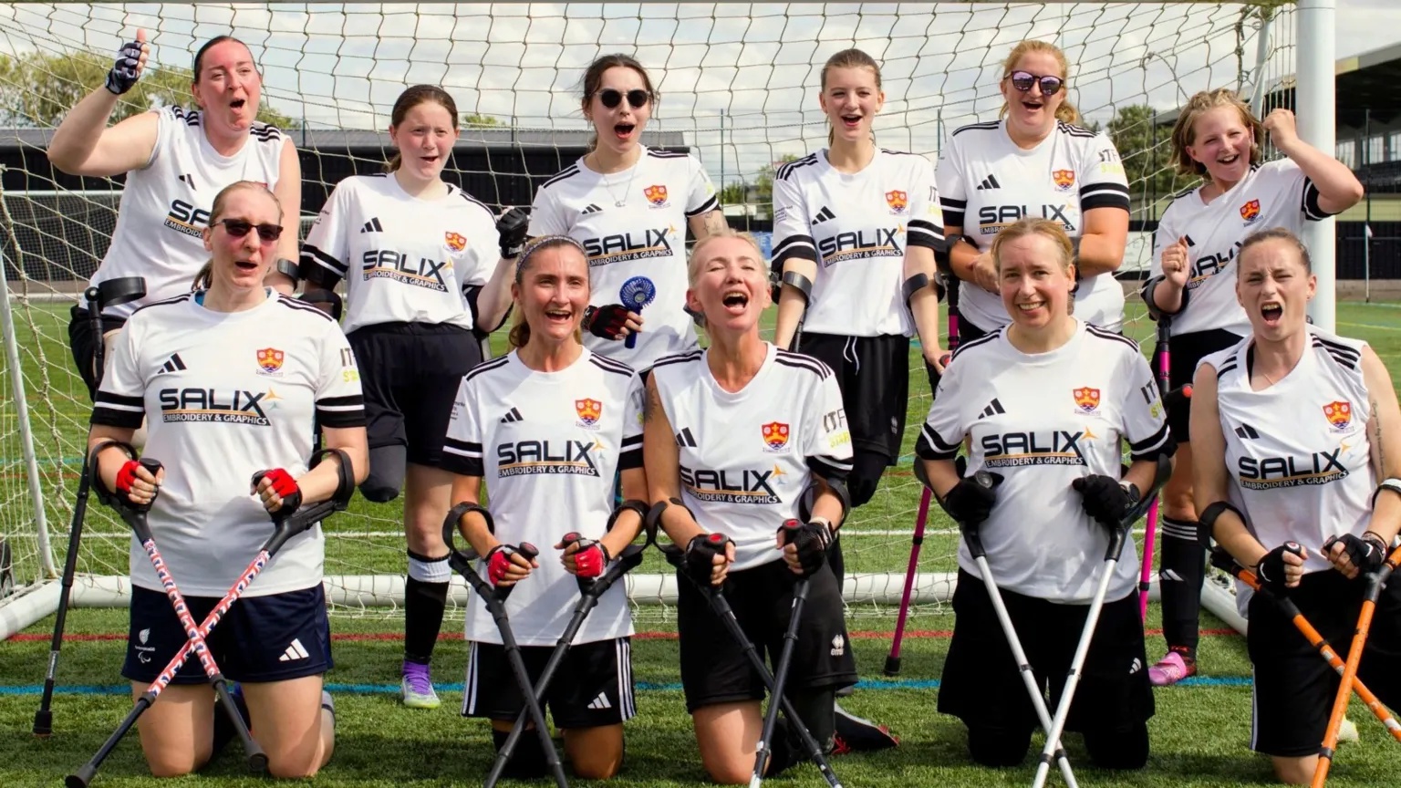 CCFC Eleven women are seen standing and kneeling in a football net at a stadium. They are all wearing white football shirts and black shorts.