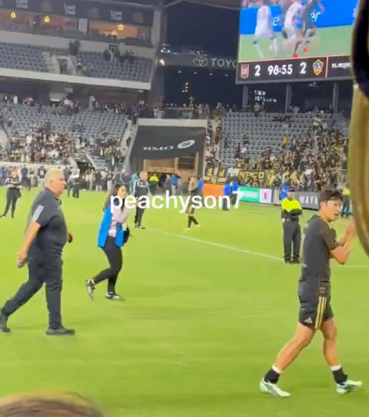 Soccer players and staff walking on the field at the end of a match at night, with the stadium scoreboard showing a tied score in the 98th minute.