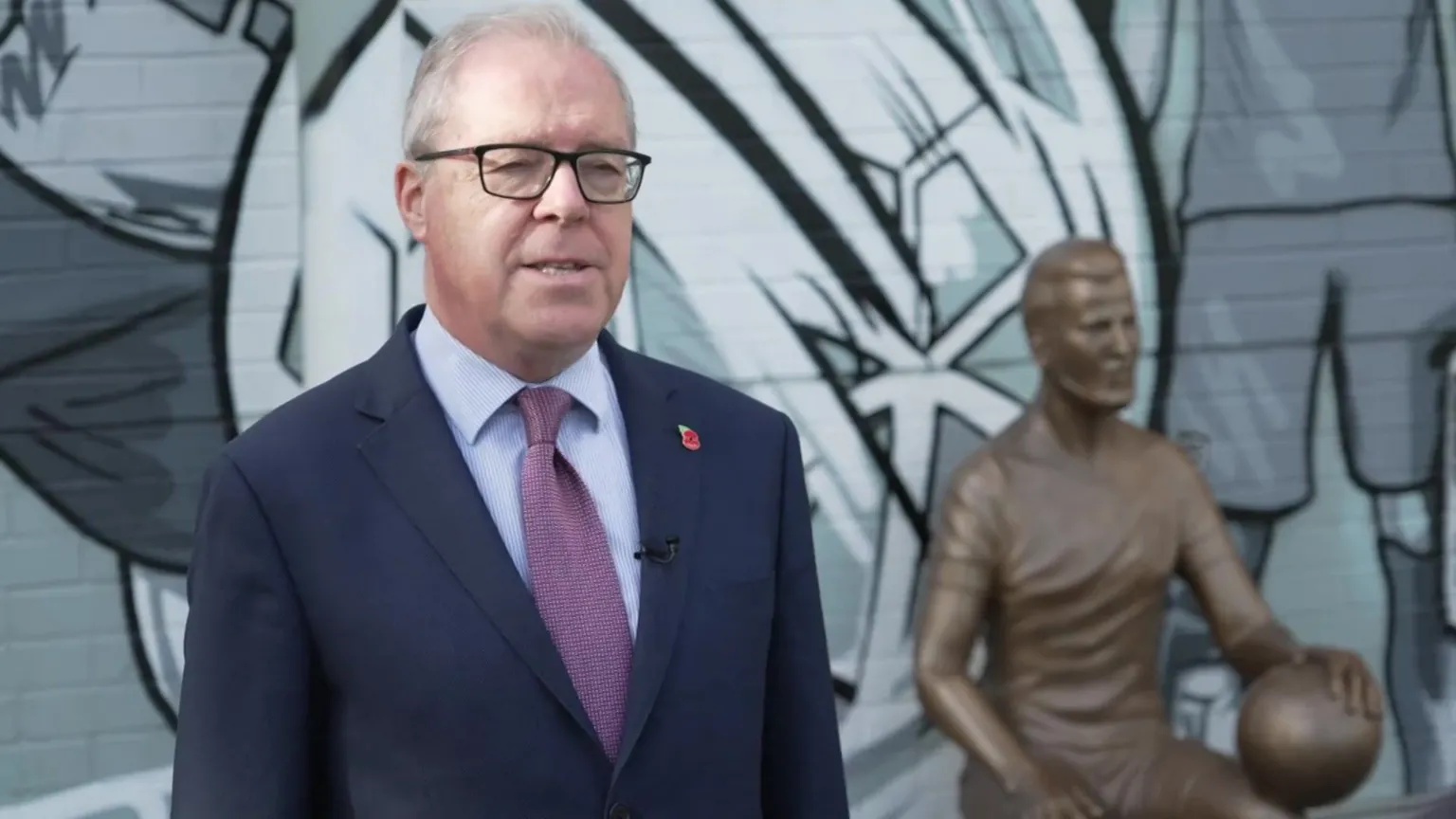 Alex Welsh in a blue suit and purple tie. He has glasses and stands in front of the Harry Kane statue.