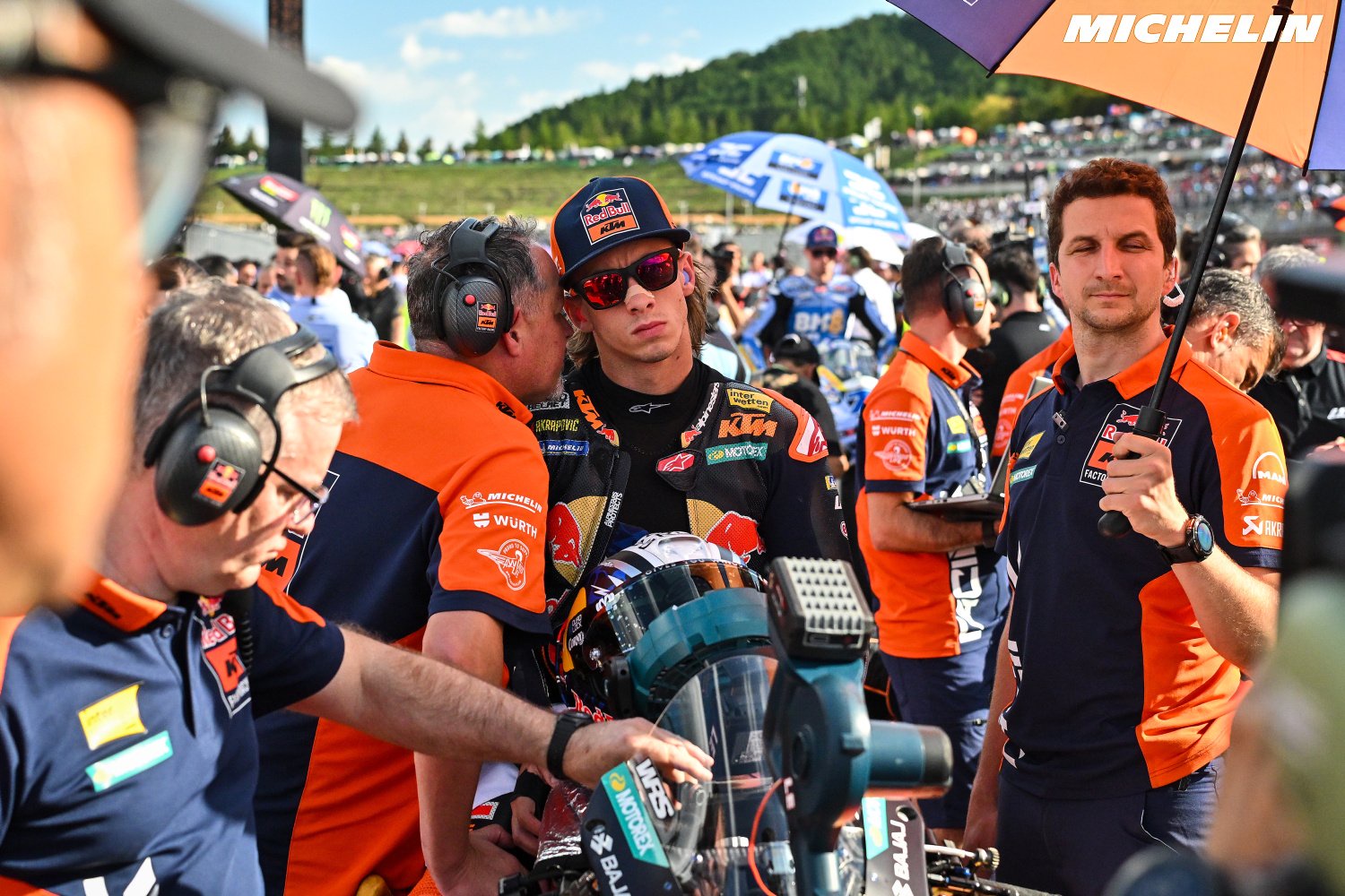 Rider in red Red Bull KTM cap and sunglasses on the MotoGP grid, surrounded by KTM team crew in orange shirts and headsets