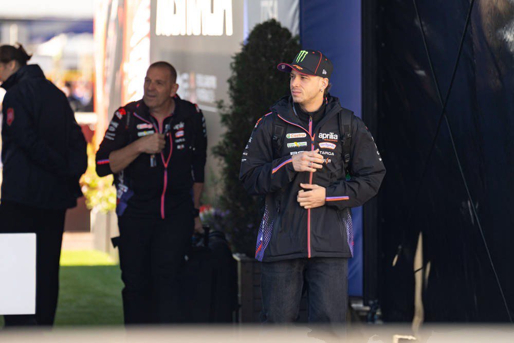 Rider in dark Aprilia team jacket with Monster cap in MotoGP paddock, standing beside a crew member in black Aprilia gear