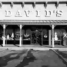 A black-and-white photo of a storefront with a sign reading "DAVID'S" above the entrance. The windows are decorated with festive holiday art, including a snowman and other winter-themed designs. There's an empty parking lot in front of the store.