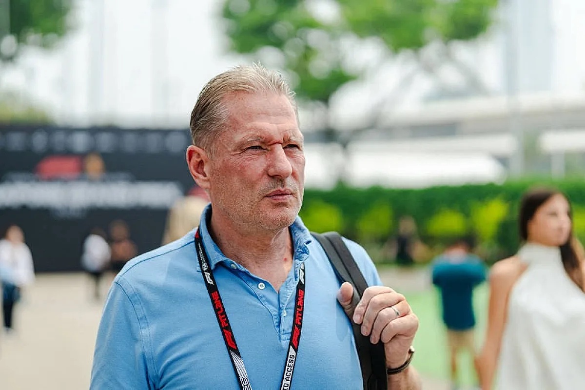 A man with grey hair in a light blue polo, wearing a lanyard, stands in a paddock area with blurred people and greenery behind him.