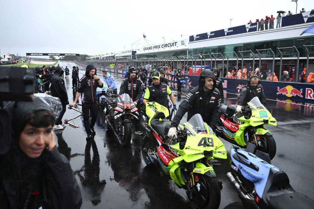 MotoGP riders in rain-soaked Yellow and black leathers push bikes on the wet grid at the Australian Grand Prix; rider #49 leads.