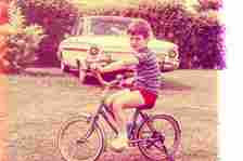 A young boy in a striped shirt and shorts rides a bicycle on grass with a vintage car parked behind him. The image has a retro aesthetic