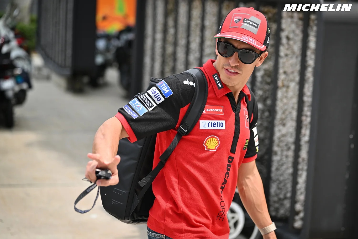 A rider in red Ducati team polo with sunglasses and cap, carrying a backpack in the MotoGP paddock, gesturing toward the camera
