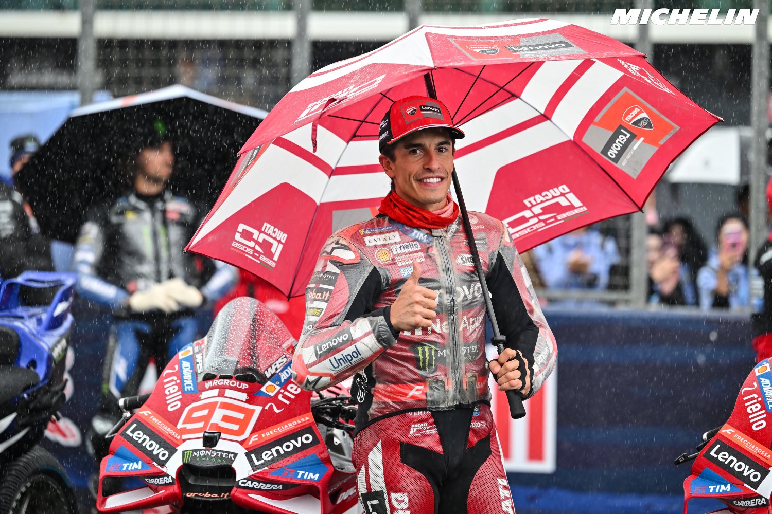 Rider in red Ducati leathers smiles under a red Ducati umbrella in the rain, giving a thumbs up beside a Ducati MotoGP bike