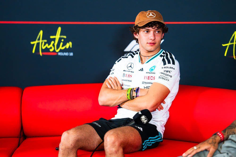 A young driver in a white Mercedes team shirt and cap sits on a red sofa with arms crossed at the Austin, Round 18 press event.