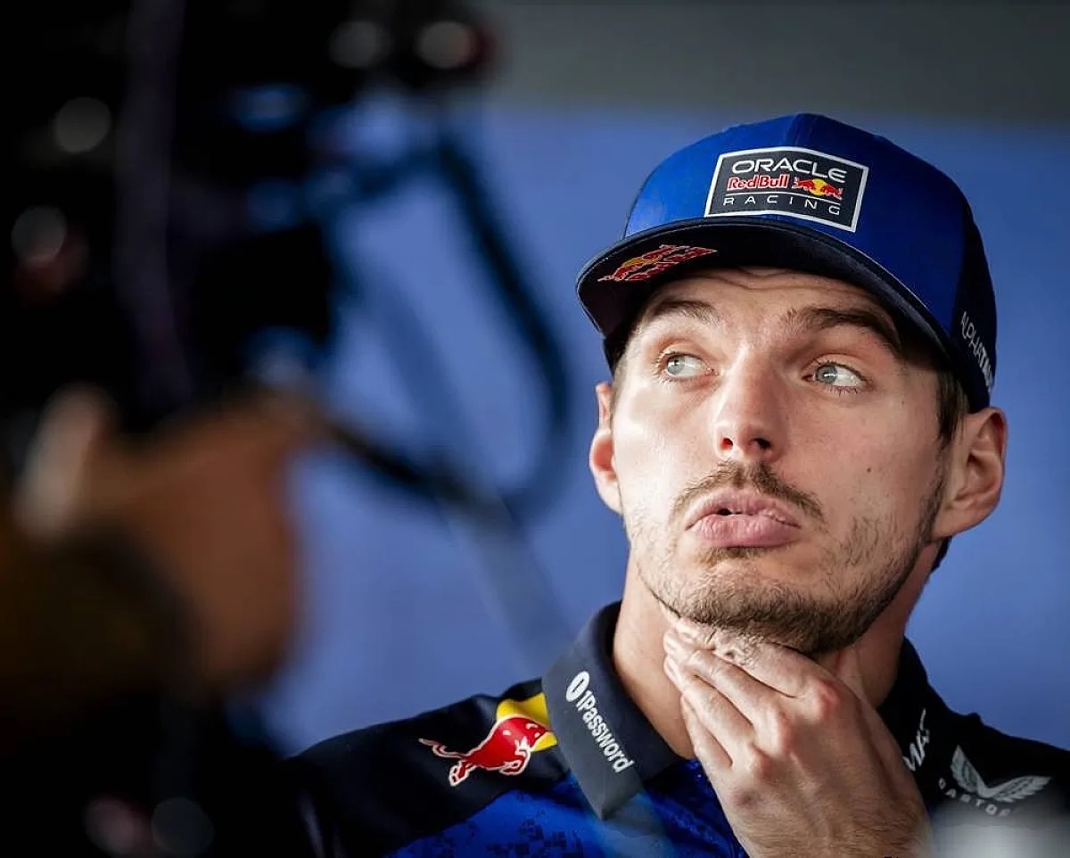 A Red Bull Racing driver in a blue cap and team shirt, looking off thoughtfully with a hand on his chin during an F1 paddock interview.