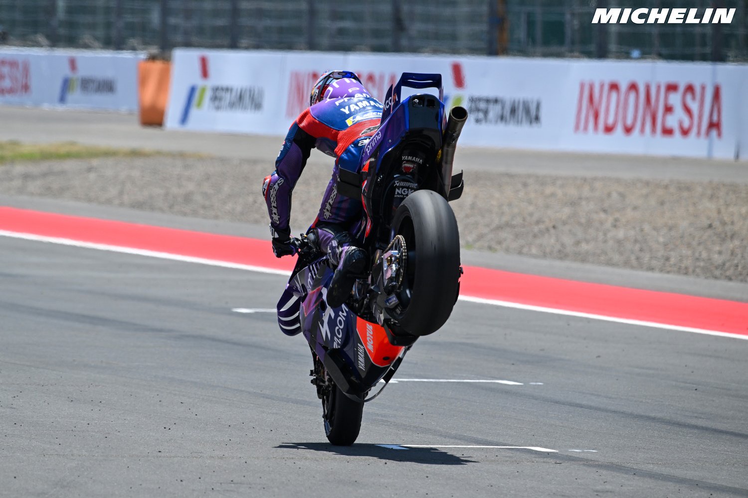 Rider in purple and red leathers performs a wheelie on a Yamaha MotoGP bike at Indonesia's track, with Pertamina banners behind.