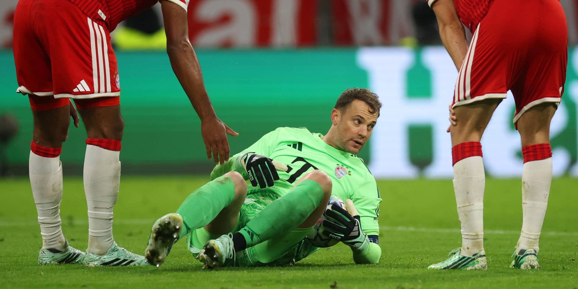 Manuel Neuer sitzt im hellgrünen Torwarttrikot am Rasen und schaut besorgt auf, während Teamkollegen neben ihm stehen.