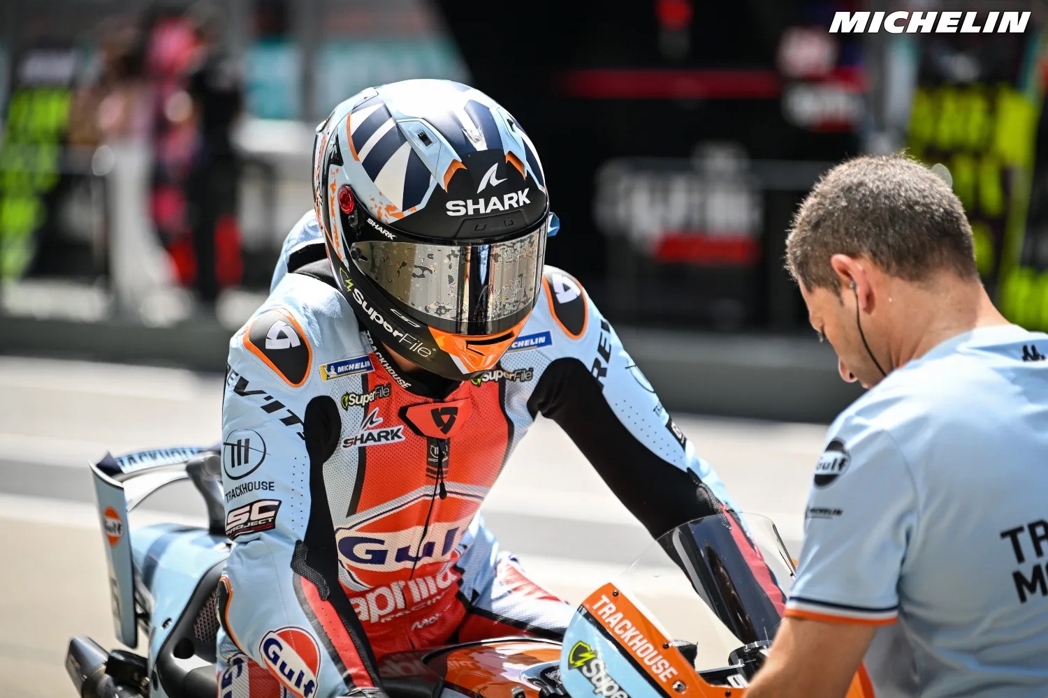 A rider in a light blue and orange Gresini-style race suit with Gulf and Shark branding, seated on a motorcycle in the pit lane, with a team mechanic beside…