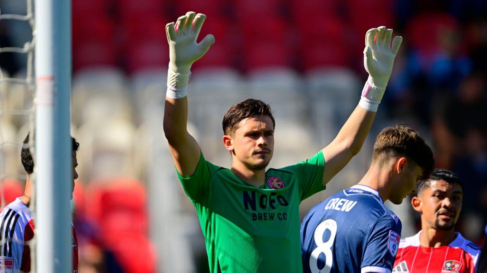 Jed Ward holding his arms in the air, wearing a green jersey and white goalkeeping gloves during a game