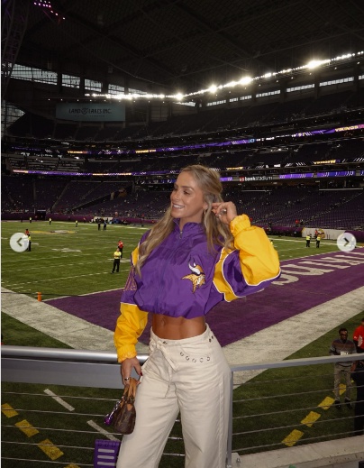 A smiling woman in a purple and yellow cropped Minnesota Vikings jacket and white pants stands in a football stadium.