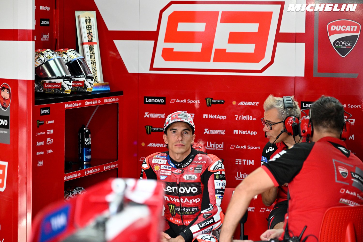 Rider in red and black Ducati leathers with number 93 sits in the Ducati Corse garage beside team staff wearing headsets, MotoGP