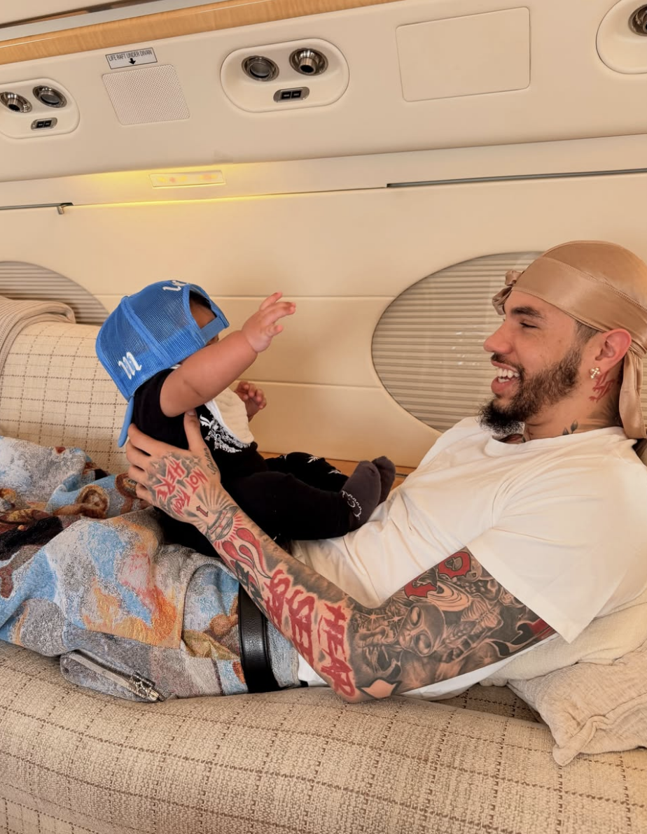 A person with tattoos and a durag smiles at a baby wearing a cap, sitting on their lap in an airplane cabin
