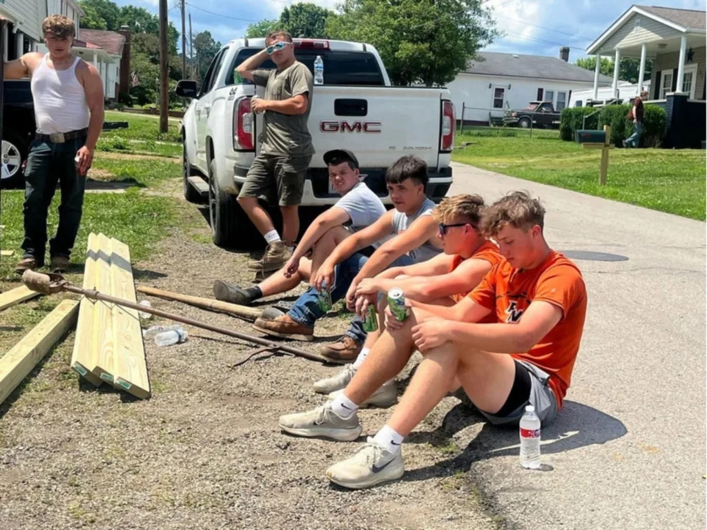 Teenage volunteers take a break on the roadside next to stacked lumber and a pickup truck
