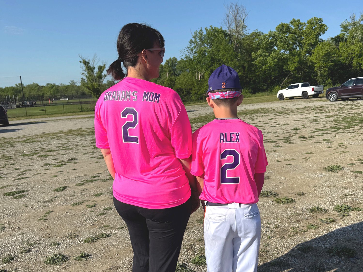 Two people stand with backs turned wearing bright pink baseball jerseys on a gravel field near parked cars.