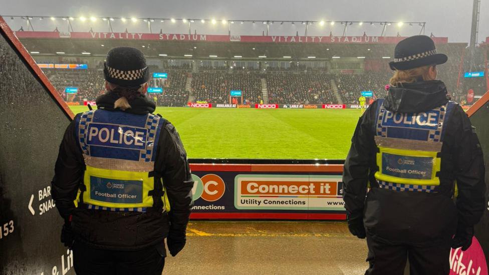 Two female police officers with their backs to the camera are looking over the pitch at the Vitality Stadium. The grass can be seen behind with a stand full of supporters. The officers are in uniform with black hats and fluorescent jackets tat say Police and Football Officer.