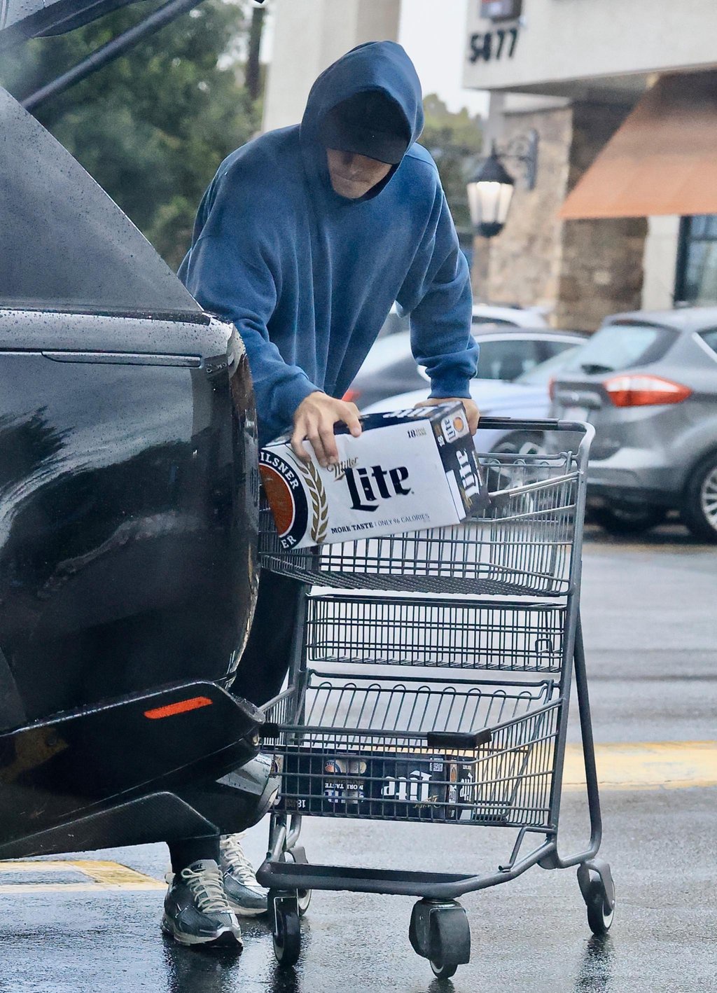 Justin Herbert wearing a blue hoodie unloads a case of Miller Lite from a shopping cart into the trunk of a car.