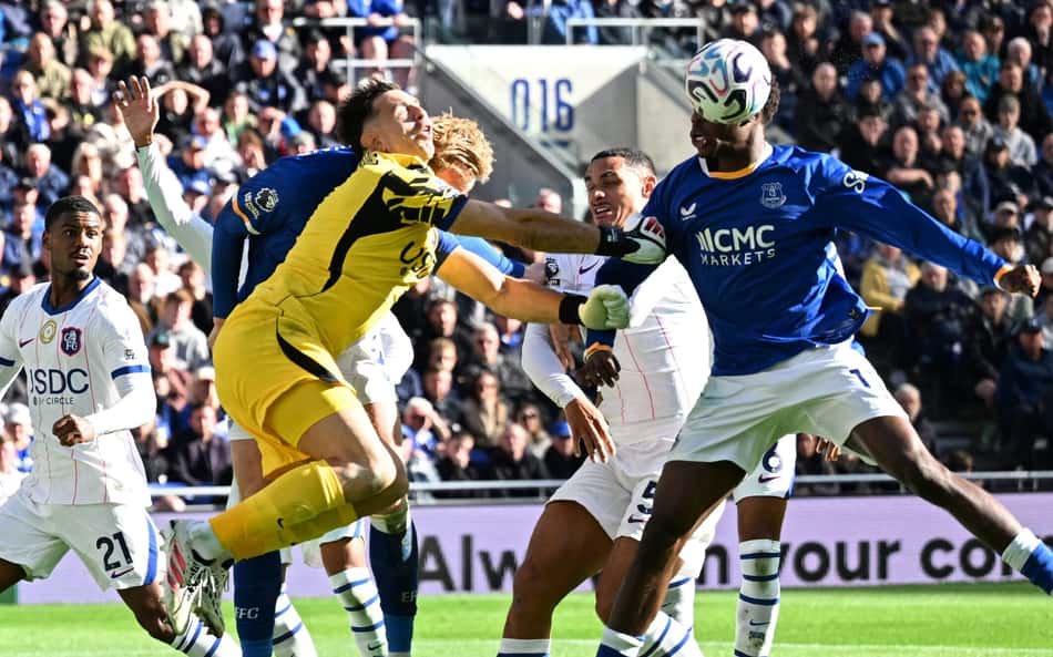 Goleiro de amarelo disputando bola com jogadores de branco e azul, em estádio com torcida ao fundo.