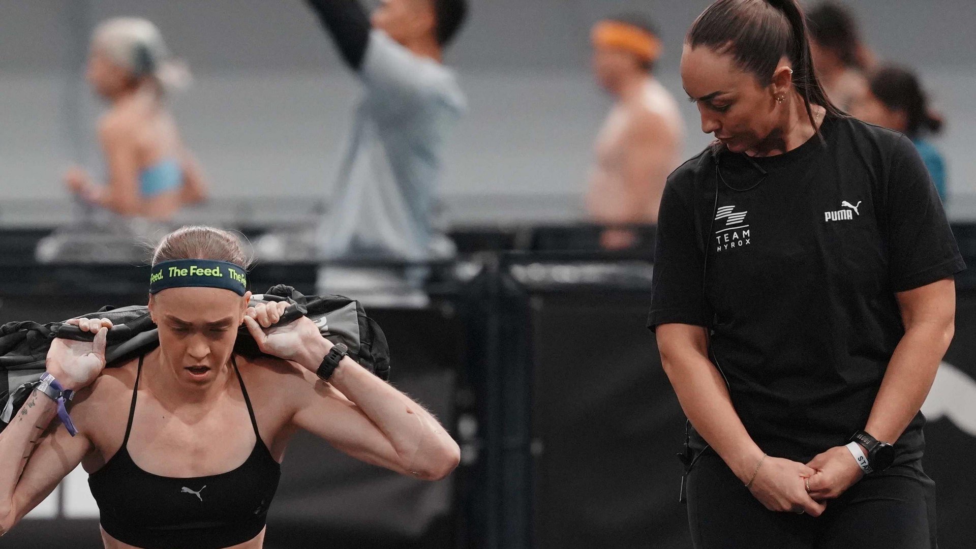 Joanna Wietrzyk competing in Hyrox competition lifting a heavy bag as an official looks on. 