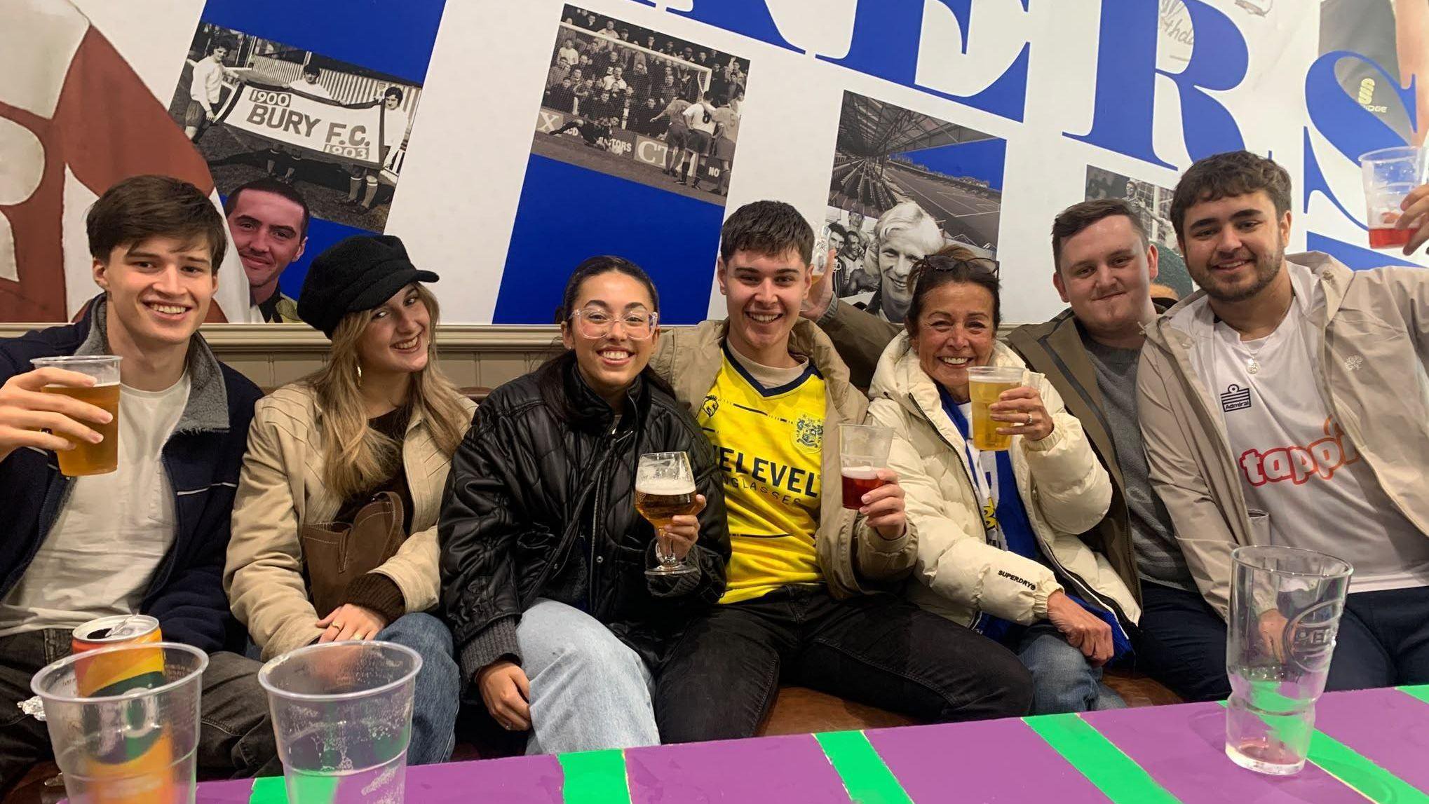 A group of Bury FC supporters sit together inside a bar area, holding drinks in front of a wall featuring historic photographs and large Bury FC-themed artwork.