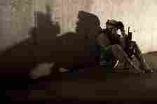 A soldier in military gear sits on the ground, head down, holding a rifle. The shadow on the wall shows the outline of Batman