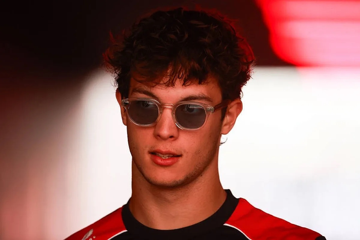 Young driver in red and black Haas-style race shirt and tinted round sunglasses in a dimly lit paddock setting