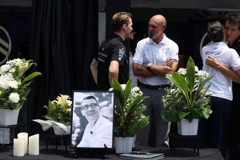 Men in team polos and a black shirt gather beside a framed black-and-white portrait with flowers and candles at a memorial tribute