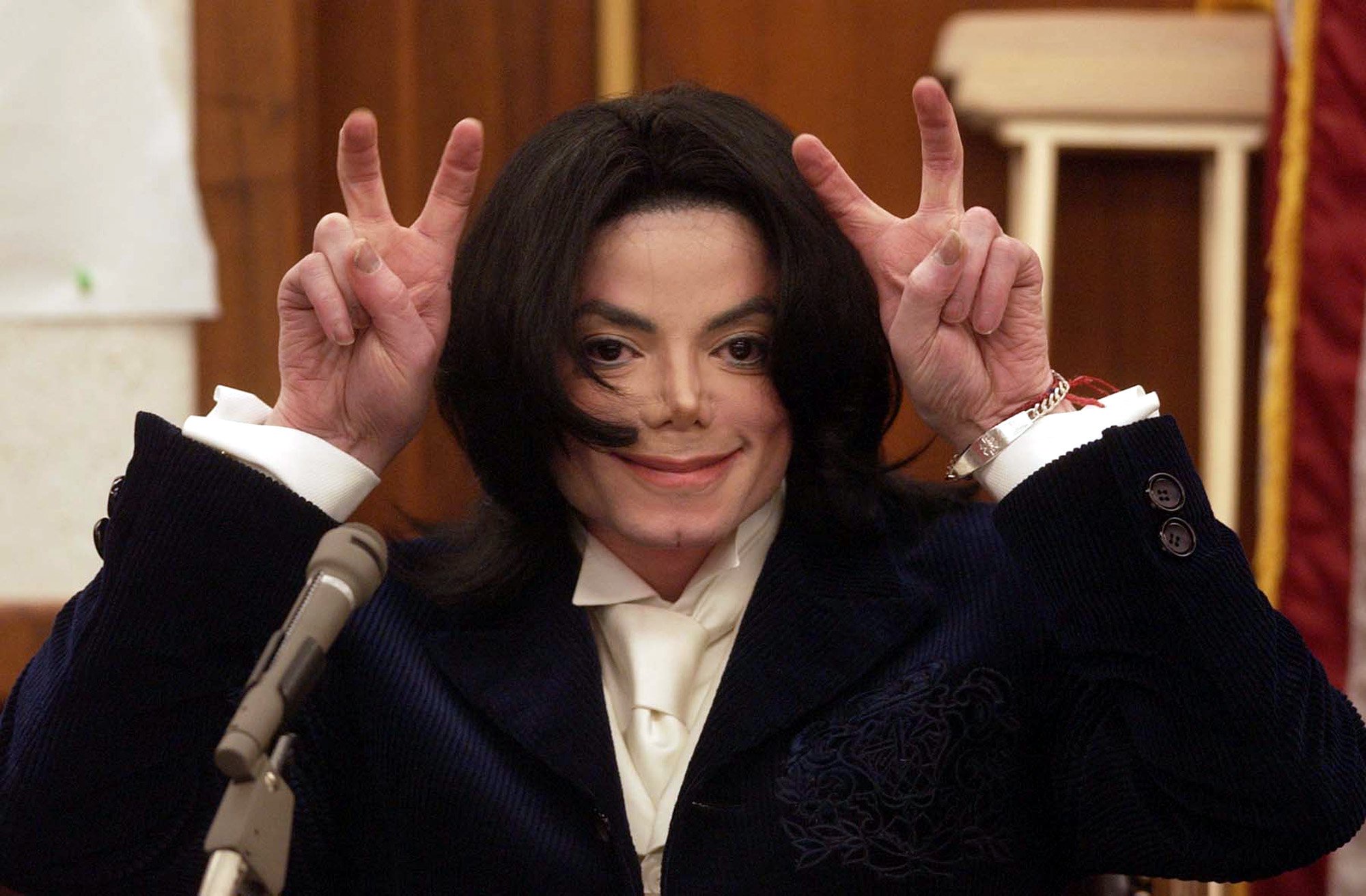 Michael Jackson making playful gestures with his hands above his head while smiling in court.