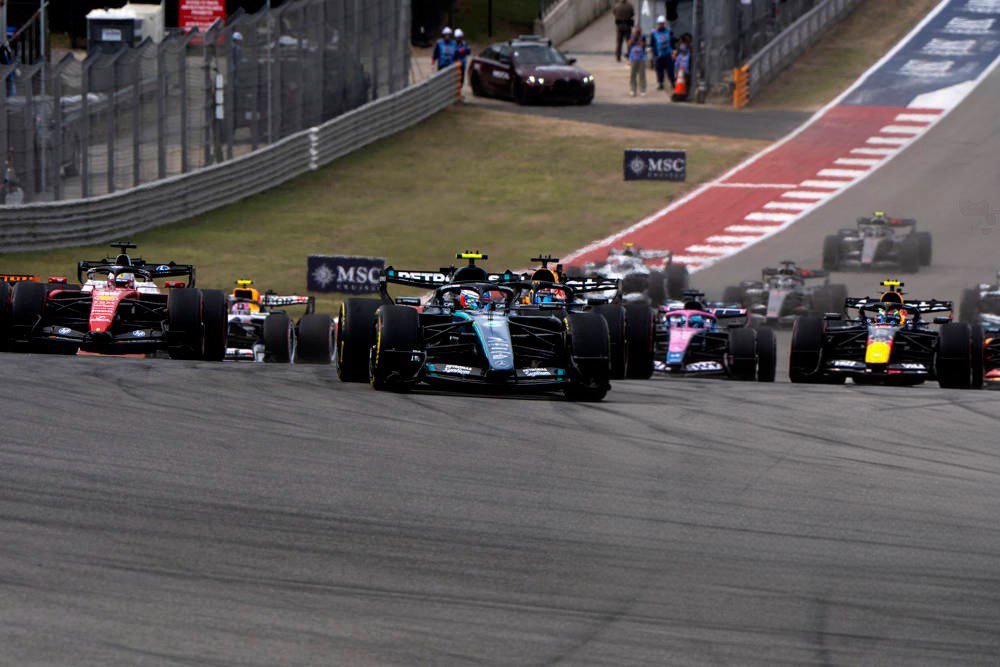 Formula 1 cars race on the Austin circuit; the lead car is a Mercedes in black and teal livery with number 12 visible, followed by a Red Bull and other teams…