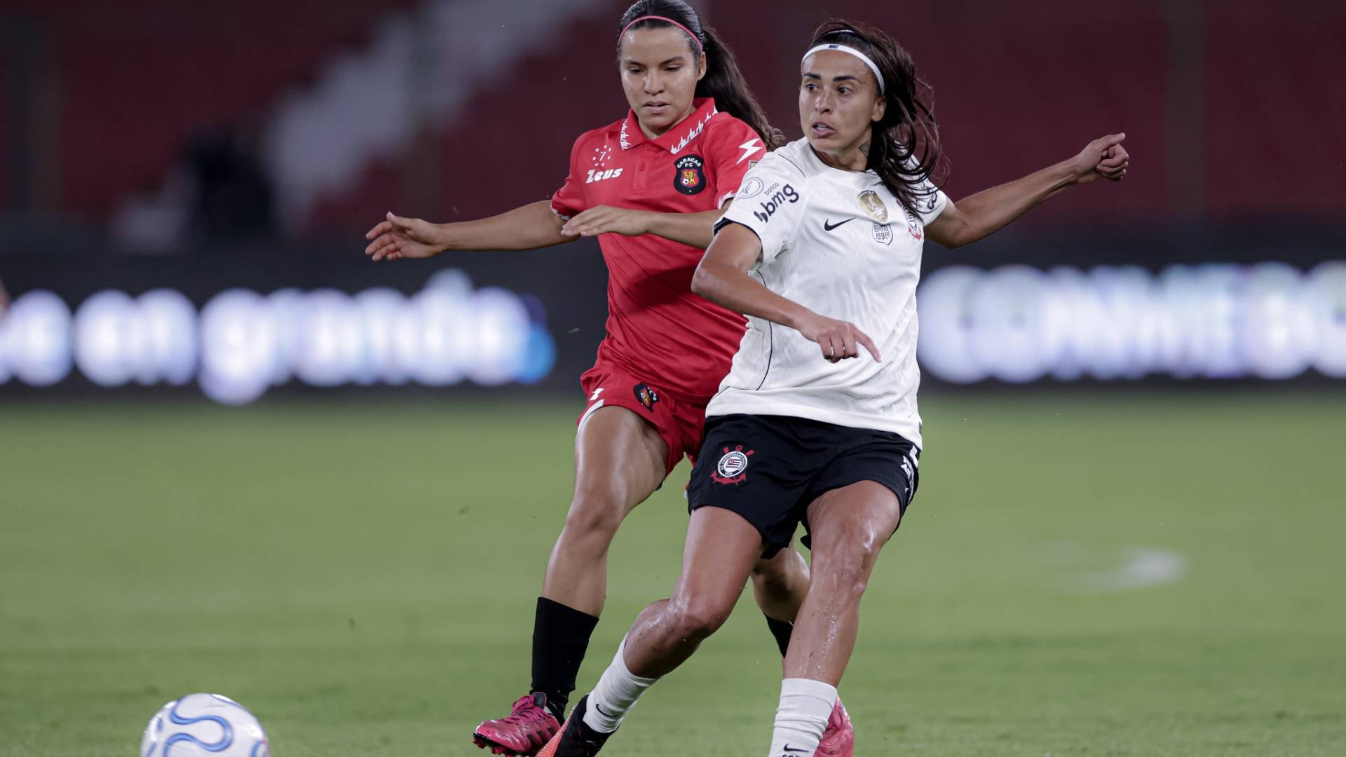 Duas jogadoras de futebol em campo verde, uma de camisa branca e shorts pretos, outra de uniforme vermelho, disputam a bola branca e azul no canto inferior esquerdo. Ao fundo, arquibancadas vermelhas e letreiros luminosos.