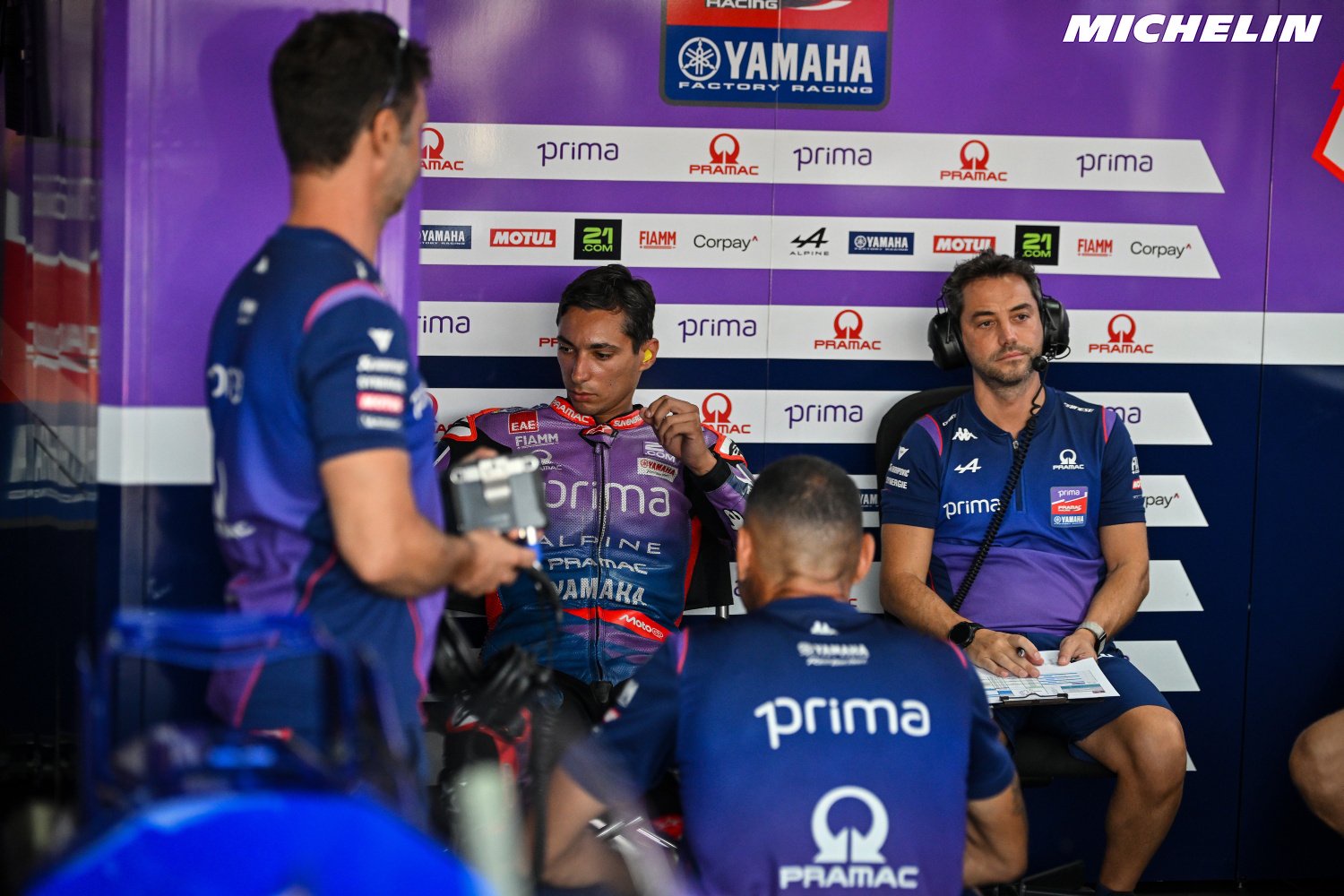 Pramac Yamaha MotoGP rider in purple-and-blue leathers sits in the team garage with headphones, while crew members stand nearby. Setting: MotoGP paddock.