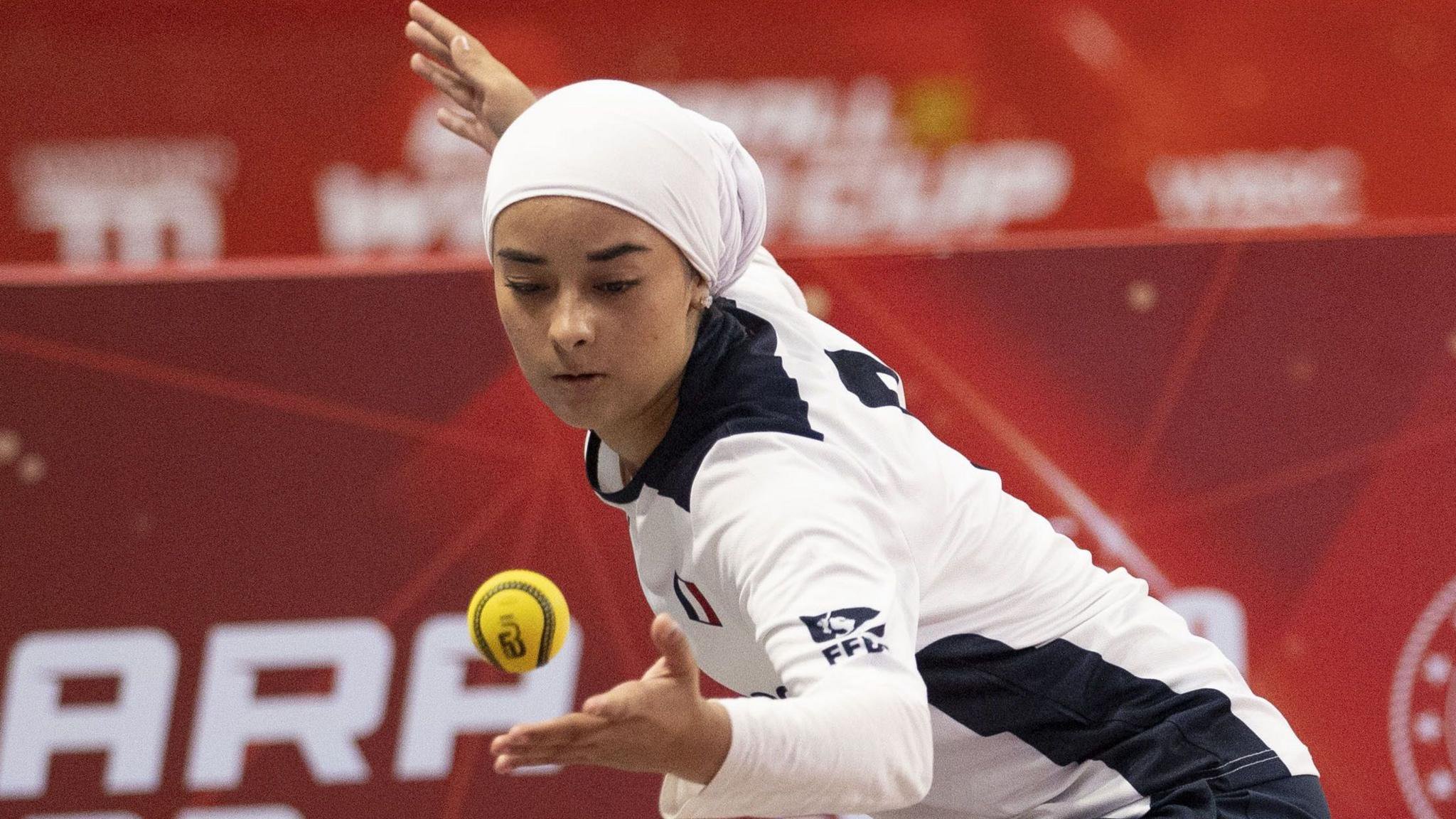 A girl wearing a white head covering and a white-and-blue long-sleeved France shirt bends forward and pulls back her right arm as she prepares to strike a yellow ball hanging in the air just about her left hand during a game of Baseball5. She stands in front of red hoardings with white writing which are out of focus