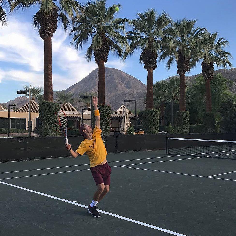 Tennis player Stefan Milicevic serving on an outdoor court with palm trees and mountains in the background.