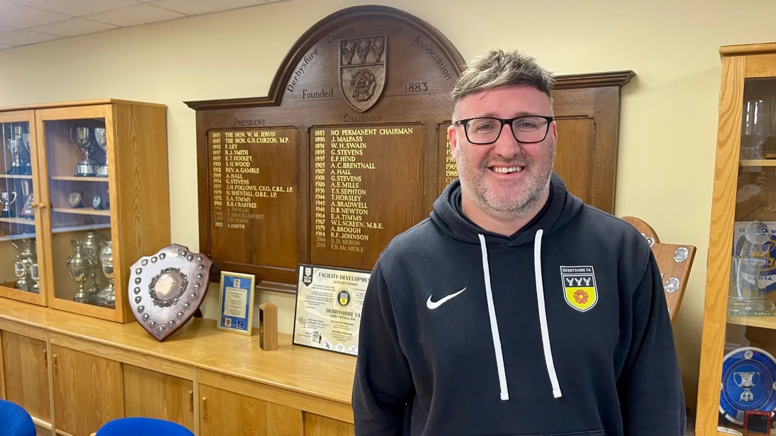 A man in a navy hoodie is stood in front of a wooden honours board in a conference room.
