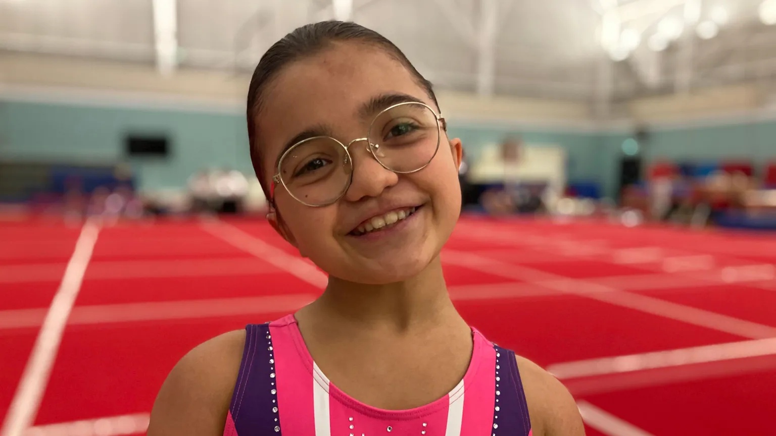 BBC Veronica smiles in a gym. She wears a pink gymnastics vest and has tied-back brown hair and glasses.