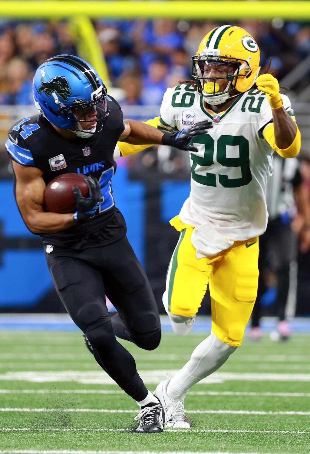 A Detroit Lions player carrying the football is pursued by a Green Bay Packers defender on the field.