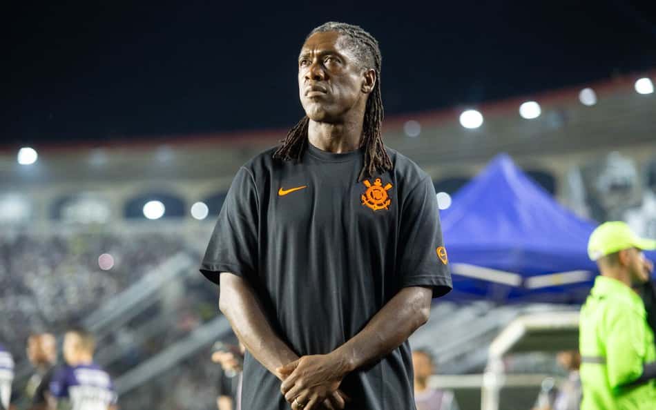 Homem com dreadlocks, camiseta preta com emblema laranja, braços cruzados em estádio durante evento.
