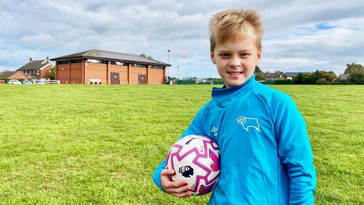 A boy in a blue tracksuit top is holding a football on a grassy field with a sports centre and floodlights in the background.