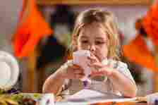 A young child is squeezing glue onto a piece of paper at a craft table, concentrating intensely on the task. Various craft materials are visible around the table